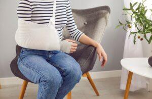 Crop unrecognizable young woman wearing a wrist bandage and an arm sling that help keep her injured limb immobile sitting on an armchair in the living room at home.